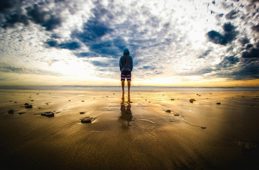 A lone figure stands on a tranquil beach during a vivid sunset, reflecting on the wet sand.